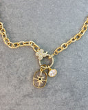 A gold-tone chain necklace with a clasp decorated with a small pavé clover and two dangling charms: a compass rose in a clear bezel and a small oval clear stone charm, displayed on a textured gray background.