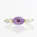 A silver-toned braided cuff bracelet featuring a central oval purple stone with a smaller faceted purple gemstone inset in the middle, accented by gold-toned bezels, set against a white background with a soft reflection.