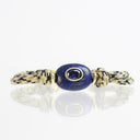 A beaded bracelet featuring a central oval lapis lazuli stone in a gold bezel setting, flanked by gold spacer beads and knotted cord ends in black and beige.