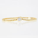A delicate yellow gold beaded bracelet featuring a single bezel-set round clear gemstone in the center, displayed on a reflective white surface.