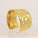 A hammered gold cuff bracelet with a textured, organic surface, displayed on a clean white reflective surface.
