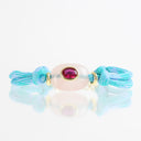 A light blue braided cord bracelet featuring a central pale pink stone bead with a gold bezel holding a round pink gemstone, set against a reflective white background.