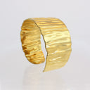 A wide, open cuff bracelet in textured yellow gold with a hammered, wavy surface finish, displayed on a clean white reflective surface.