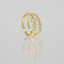 A gold ring with a double-band design, featuring a row of small clear gemstones on one band and a textured beaded pattern on the other, displayed on a reflective light gray surface.