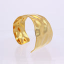 A hammered gold cuff bracelet with a unique organic wavy shape, displayed on a clean white reflective surface.
