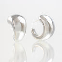 A pair of polished silver crescent-shaped hoop earrings shown from two angles on a reflective white surface.