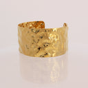 A hammered gold cuff bracelet with a textured, organic surface, displayed on a reflective white surface against a light background.
