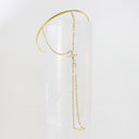 A delicate gold chain necklace with a small cross pendant draped over a clear cylindrical display stand against a white background.