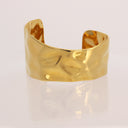 A polished gold cuff bracelet with a textured, hammered finish, displayed on a clean white reflective surface.
