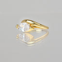 A yellow gold ring featuring an oval-cut clear gemstone in a prong setting, displayed on a reflective light gray surface.