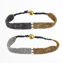Two woven chain bracelets shown on a white background, one in black and gold and one in silver and gold, each with a braided black cord and a round gold bead clasp.