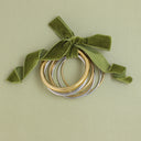 A set of three stacked metal bangles in gold, silver, and rose gold, tied with an olive green velvet ribbon bow, on a solid sage green background.
