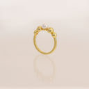 A gold ring featuring a polished band and three small gold beads at the top, with a single white pearl centered on the front, set against a soft cream background with a subtle reflection.