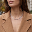 A close-up lifestyle shot of a woman wearing a delicate silver tennis necklace with clear stones, styled with a camel-colored wool coat.