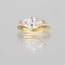 A yellow gold ring featuring a large marquise-cut clear gemstone in a prong setting, displayed on a reflective light gray surface.