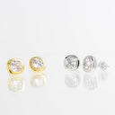 Two pairs of cushion-cut diamond stud earrings shown on a reflective white surface. One pair features a yellow gold bezel setting, and the other pair features a silver-toned bezel setting.