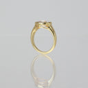A yellow gold ring with a bezel-set clear gemstone, shown from a side profile view on a reflective light gray surface.