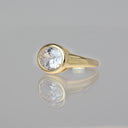 A yellow gold ring featuring a large, round brilliant-cut clear gemstone in a bezel setting, displayed on a reflective gray surface.