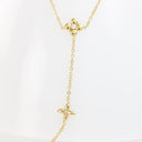 A delicate gold lariat necklace featuring two small four-petal flower charms, each set with a clear round gemstone, displayed on a white background.