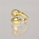 A gold ring with a teardrop-shaped bezel holding a clear round gemstone, displayed on a reflective light gray surface.
