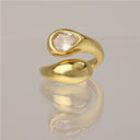 A polished gold ring with a teardrop-shaped clear gemstone set in a curved, open design, displayed on a reflective light gray surface.