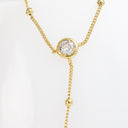 A gold Y-necklace featuring a central bezel-set round clear gemstone, with delicate chain drapes and small gold bead accents on a white background.