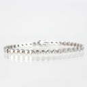 A silver tennis bracelet with a continuous line of polished round links and a clasp, displayed on a reflective white surface against a white background.