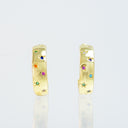 A pair of gold huggie hoop earrings decorated with small colorful gemstones, shown on a light gray reflective background.