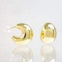 A pair of shiny gold crescent moon stud earrings on a reflective white surface, showing one earring from the side with its clear post and the other from the back.