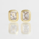 A pair of gold rectangular stud earrings with clear rectangular-cut gemstones, displayed on a reflective white surface.