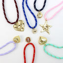 A collection of colorful braided cord necklaces with gold-toned charms, arranged on a clean white background. The necklaces feature heart, star, shell, and flower charms in various colors and finishes, paired with cords in burgundy, blue, navy, pink, lavender, red, and mint green.