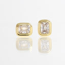 A pair of gold stud earrings with rectangular cushion-cut clear gemstones, displayed on a reflective white surface.
