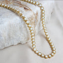 A gold chain necklace with round white pearl-like beads draped over a textured white stone prop on a light beige fabric background.