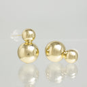 A pair of gold double-ball stud earrings with a clear silicone backing, displayed on a reflective light gray surface.