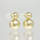 A pair of gold double-ball stud earrings with clear plastic backing, displayed on a reflective light gray surface.