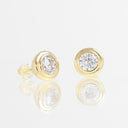 A pair of gold stud earrings with round bezel-set clear gemstones, displayed on a reflective white surface.