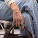 A gold chain hand bracelet with small light blue gemstone accents worn on a person's hand, shown while they sit on a white woven chair and hold a brown leather handbag.