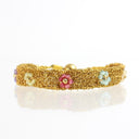 A gold mesh bracelet decorated with small colorful enamel flowers in pink, light blue, and pale yellow, shown on a reflective white surface.