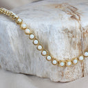 A gold tennis necklace with round white opal-like inlays, draped over a textured light gray stone slab.
