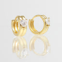 A pair of gold huggie hoop earrings, each featuring a small clear heart-shaped gemstone in a prong setting, displayed on a reflective white surface.