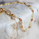 A gold chain necklace with marquise-cut red gemstones and small clear stones, draped over a textured white stone slab.