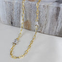 A gold paperclip chain necklace with two silver barbed wire charms, displayed on a light beige fabric surface with a rustic gray wooden block in the background.