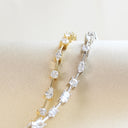 Two tennis-style bracelets with clear rectangular and marquise-cut stones, one in yellow gold and one in silver-tone metal, displayed on a cream fabric surface.