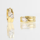 A pair of gold huggie hoop earrings, each featuring a marquise-cut clear gemstone in a prong setting. The earrings are displayed on a reflective white surface against a clean white background.