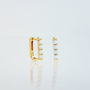 A pair of gold huggie hoop earrings with a row of white opal gemstones, displayed on a reflective white surface against a light gray background.