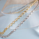 Two teardrop-link necklaces with clear gemstones, one in yellow gold and one in silver, displayed on a clear acrylic stand against a light blue background.