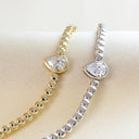 Two beaded chain bracelets displayed on a cream fabric surface, one in yellow gold and one in silver, each featuring a pear-cut clear gemstone centerpiece.
