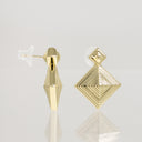 A pair of gold geometric stud earrings with a faceted diamond design, one shown from the front and the other from the side with a clear silicone earbud tip, on a light gray reflective surface.