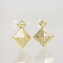 A pair of gold geometric stud earrings with a textured, concentric diamond design and clear butterfly backings, displayed on a reflective light gray surface.