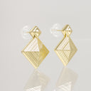 A pair of gold geometric stud earrings with a faceted diamond-shaped drop and a textured, ribbed surface, shown on a light gray reflective surface.
